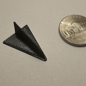 Puede incluir: Un pequeño modelo de avión de papel negro impreso en 3D con una superficie texturizada. Junto a él hay una moneda estadounidense de cinco centavos. El modelo mide aproximadamente 2,5 cm de largo.