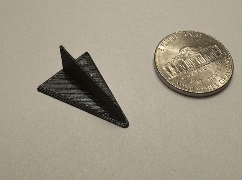 K&ouml;nnte beinhalten: Ein kleines, schwarzes, 3D-gedrucktes Papiermodell eines Flugzeugs mit strukturierter Oberfl&auml;che. Daneben liegt eine amerikanische F&uuml;nf-Cent-M&uuml;nze. Das Modell ist etwa 2,5 cm lang.