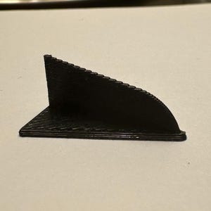 K&ouml;nnte beinhalten: Kleines, schwarzes, 3D-gedrucktes Objekt mit dreieckiger Basis und gebogenem, geriffeltem Oberteil. Das Objekt scheint eine St&uuml;tze oder ein dekoratives Element mit strukturierter Oberfl&auml;che zu sein. Das Design ist minimalistisch und modern.