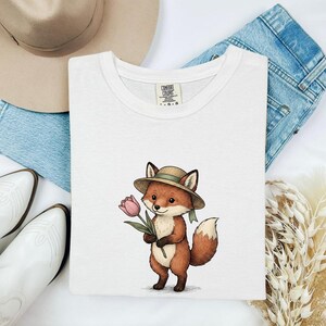 Puede incluir: Una camiseta blanca de cuello redondo con un gráfico de un zorro de dibujos animados que lleva un sombrero de paja y sostiene un tulipán rosa. La prenda tiene una etiqueta de la marca Comfort Colors en el cuello. El diseño está centrado.