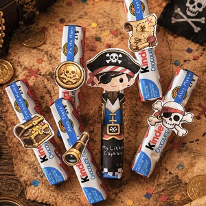 May include: Five Kinder chocolate bars decorated with pirate-themed paper cutouts. The cutouts feature a treasure chest, a gold coin, a pirate character labeled My Little Captain, a treasure map, and a skull with an eyepatch. Blue paper bands include text like Yo Ho Hero, Ahoy There, and Treasure Hunter.