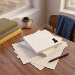May include: A stack of square white paper sheets featuring a small black silhouette of a corgi dog in the corner. The paper is layered on top of larger, light-colored textured sheets, accompanied by a black pen on a wooden surface.