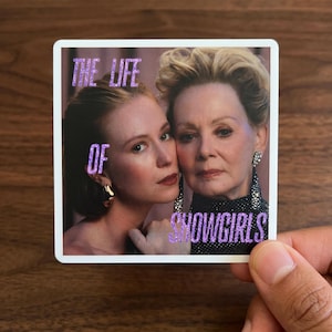 May include: A square sticker featuring a photographic portrait of two individuals with blonde hair, one wearing a gold earring and the other wearing a black textured top with long silver earrings. The text THE LIFE OF SHOWGIRLS is in purple.