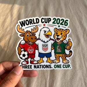 May include: A die-cut sticker featuring the text WORLD CUP 2026 and THREE NATIONS. ONE CUP. The design shows a cartoon moose in a red Canada jersey, a bald eagle in a white USA jersey, and a jaguar in a green Mexico jersey holding a soccer ball.
