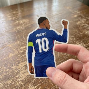 May include: A die-cut sticker featuring a soccer player seen from behind, wearing a dark blue long-sleeve jersey with the name MBAPPÉ and the number 10 printed in white on the back, along with a bright yellow armband on the left sleeve.