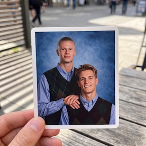 May include: A rectangular photograph showing two individuals wearing light blue button-down shirts layered under dark green and black argyle sweater vests. The subjects are positioned against a mottled blue backdrop in a classic portrait style.