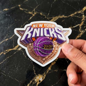 May include: A die-cut sticker featuring the New York Knicks logo in purple, orange, and white. The design includes a basketball with intricate filigree patterns, the text New York Knicks 2017, and a metallic-toned shield background.