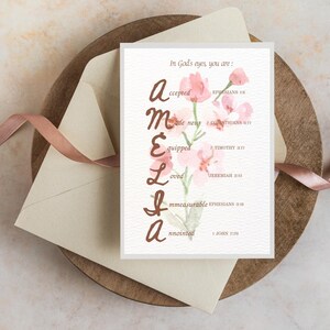 May include: A greeting card with the name "Amelia" spelled out vertically with words describing her. The card features a watercolor floral design in pink and green. Text on the card reads "In God's eyes, you are: accepted, made new, equipped, loved, immeasurable, anointed."