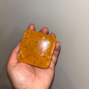 May include: A square-shaped bar of soap with a smooth, glossy finish. The soap is a vibrant orange color and features numerous small, dark brown speckles distributed throughout the surface, giving it a textured appearance within the solid form.
