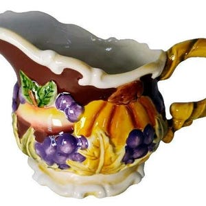 May include: A ceramic creamer jug featuring a brown background with a raised, three-dimensional relief design of yellow pumpkins, green leaves, and clusters of purple grapes. The jug has a scalloped rim and a twisted, yellow-toned handle.