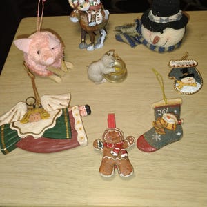 May include: A collection of eight holiday ornaments including a pink pig, a Santa on a reindeer, a snowman head with a blue scarf, a cat with a bowl, a mitten with a snowman, an angel, a gingerbread person, and a stocking with a snowman and the word JOY.