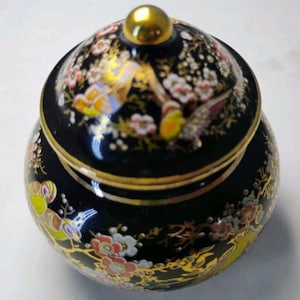 May include: A small, round, black ceramic jar with a matching lid featuring a gold-colored knob. The surface is decorated with a colorful floral pattern in shades of red, white, and yellow, accented with gold-colored branches and bird motifs.
