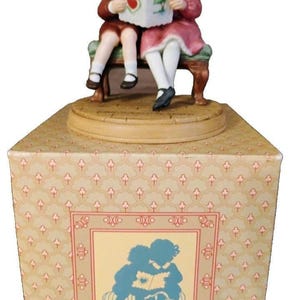 May include: A porcelain figurine of two children sitting on a bench reading a book, placed atop a square box. The box features a repeating floral pattern and a central graphic of the same two children in silhouette, rendered in blue and cream.
