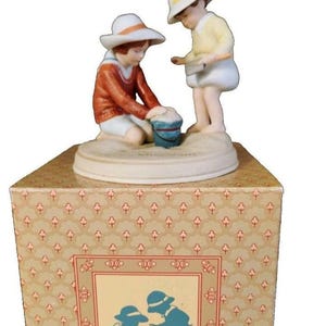 May include: A porcelain figurine of two children wearing white sun hats and playing in the sand with a blue bucket. One child wears a rust-colored sweater and white shorts, while the other wears a yellow sweater and white shorts. The base is circular.