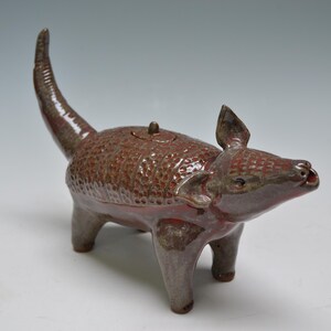 May include: A brown ceramic teapot shaped like an armadillo with a glossy finish. The teapot features a textured, segmented shell pattern on its back, a long upright tail, four sturdy legs, pointed ears, and a small lid centered on its back.