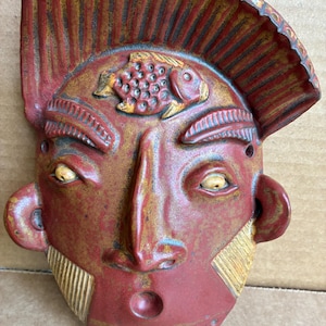 May include: A red and yellow ceramic wall mask featuring a stylized face with a prominent forehead crest, a fish relief on the forehead, textured eyebrows, round eyes, large ears, and diagonal etched lines on the cheeks and chin area.