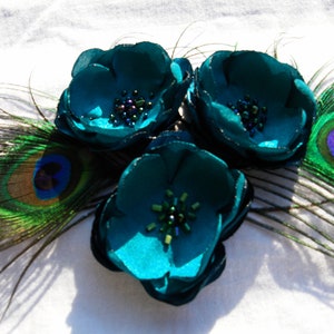 May include: A teal floral hair accessory featuring three fabric flowers with beaded centers. Peacock feathers in shades of green, blue, and gold are arranged around the flowers, creating a decorative accent.