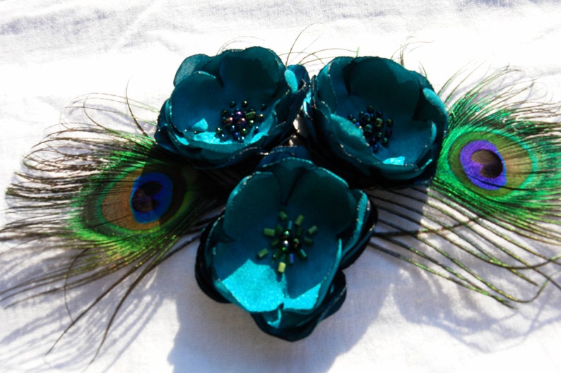 May include: A teal floral hair accessory featuring three fabric flowers with beaded centers. Peacock feathers in shades of green, blue, and gold are arranged around the flowers, creating a decorative accent.