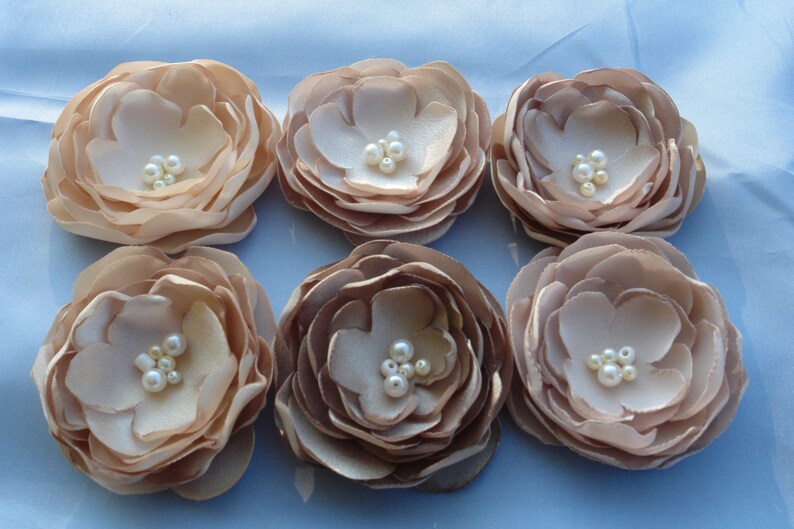 May include: Six beige fabric flowers with pearl embellishments. The flowers are arranged in two rows of three.