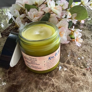 Könnte beinhalten: Ein Glasgefäß mit einem gelb-grünen Balsam, einem schwarzen Deckel und einem Etikett mit der Aufschrift King Of Balm. Das Etikett führt Inhaltsstoffe wie Ringelblume, Rosmarin und Steinklee auf sowie Text zur Linderung von Muskelbeschwerden.