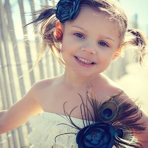 May include: A young girl wearing a white lace dress and a teal flower hair accessory with peacock feathers. The dress has a strapless design and is adorned with a teal flower and peacock feathers. The image has the text "Sweet Magnolia Photography - NY".