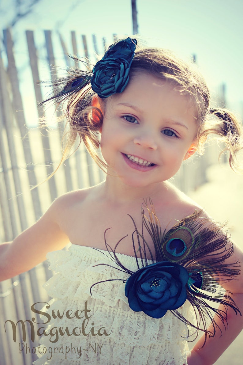 May include: A young girl wearing a white lace dress and a teal flower hair accessory with peacock feathers. The dress has a strapless design and is adorned with a teal flower and peacock feathers. The image has the text "Sweet Magnolia Photography - NY".