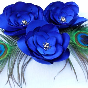 May include: Three blue satin flowers with pearl accents, surrounded by two peacock feathers with green and blue eyes.