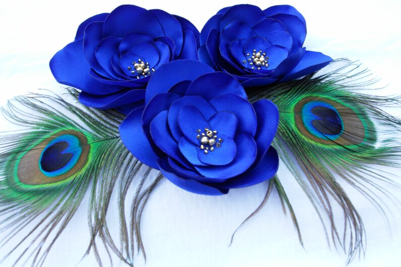 May include: Three blue satin flowers with pearl accents, surrounded by two peacock feathers with green and blue eyes.