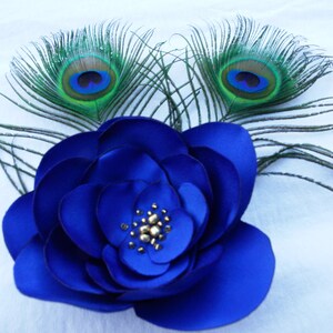 May include: A blue fabric flower with a gold center, accented with two peacock feathers with green and blue eyes.