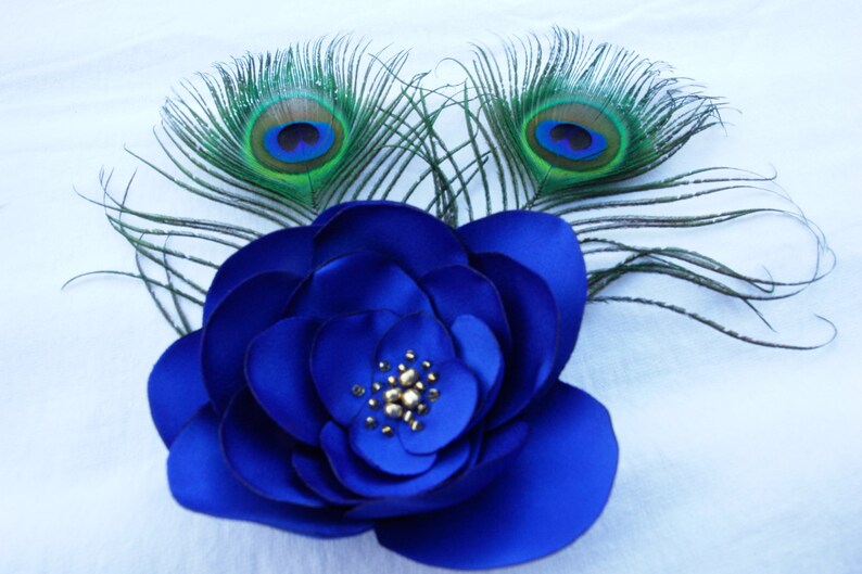 May include: A blue fabric flower with a gold center, accented with two peacock feathers with green and blue eyes.