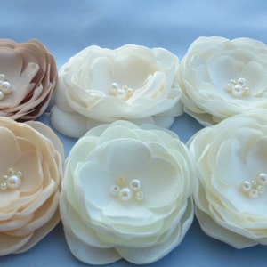 May include: Six fabric flowers with pearl embellishments. The flowers are in shades of ivory and beige.