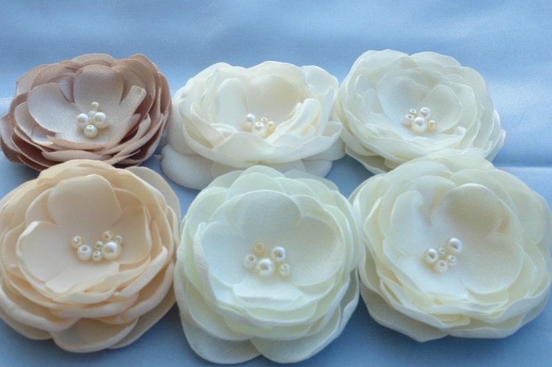 May include: Six fabric flowers with pearl embellishments. The flowers are in shades of ivory and beige.