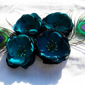 May include: A cluster of four teal fabric flowers with beaded centers and shimmering edges, accented by two peacock feathers. The flowers are arranged on a white surface, creating a striking contrast of colors and textures.