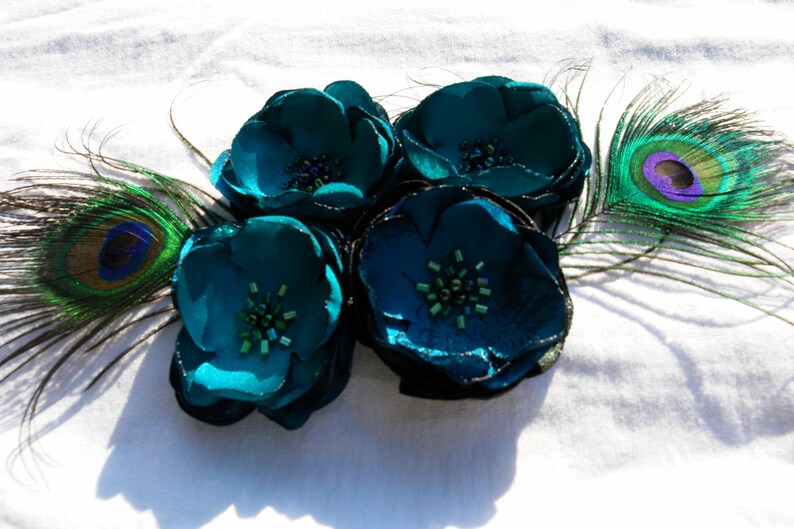May include: A cluster of four teal fabric flowers with beaded centers and shimmering edges, accented by two peacock feathers. The flowers are arranged on a white surface, creating a striking contrast of colors and textures.