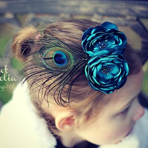 May include: A teal floral hair clip with a peacock feather accent. The clip features two fabric flowers and a single peacock feather. The image includes the text "Sweet Magnolia Photography-NY".