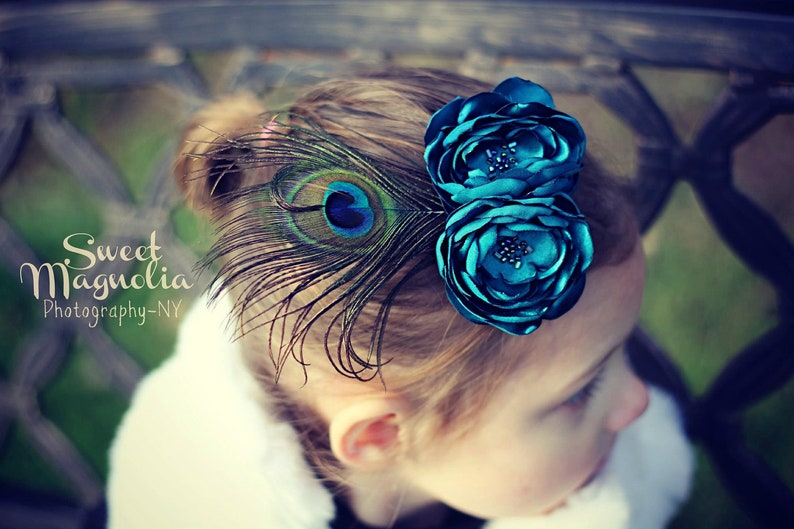 May include: A teal floral hair clip with a peacock feather accent. The clip features two fabric flowers and a single peacock feather. The image includes the text "Sweet Magnolia Photography-NY".