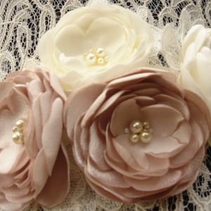 May include: A white lace fabric sash with a flower embellishment. The flower embellishment is made of three large fabric flowers, two are a light brown color and one is white. The flowers are accented with pearl-like beads.