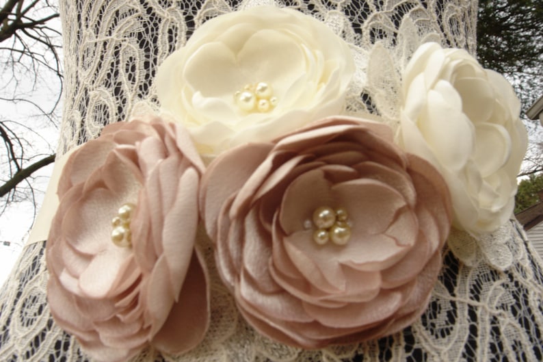 May include: A white lace fabric sash with a flower embellishment. The flower embellishment is made of three large fabric flowers, two are a light brown color and one is white. The flowers are accented with pearl-like beads.