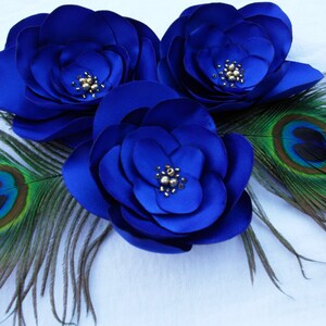 May include: Three large blue satin flowers with gold bead centers, accented by two peacock feathers with green and blue eyes.