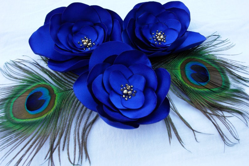 May include: Three large blue satin flowers with gold bead centers, accented by two peacock feathers with green and blue eyes.