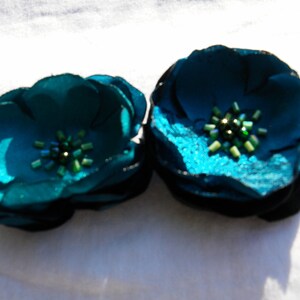 May include: Two teal fabric flower hair clips. Each flower has multiple petals and a cluster of green beads in the center. The flowers are on a white background.
