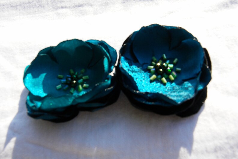 May include: Two teal fabric flower hair clips. Each flower has multiple petals and a cluster of green beads in the center. The flowers are on a white background.
