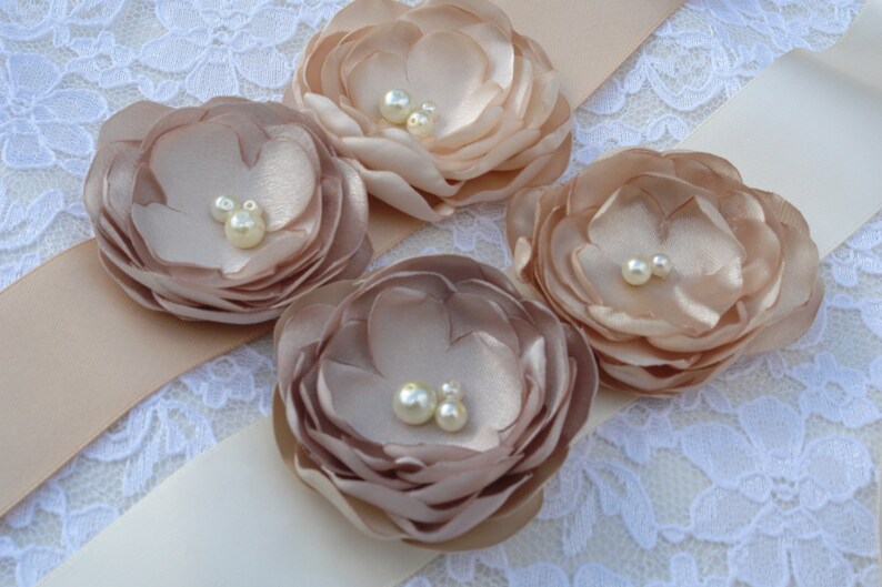 May include: A satin ribbon sash with four blush-colored fabric flowers, each with pearl embellishments.