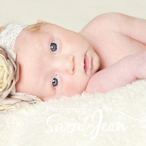 May include: A newborn baby girl wearing a white lace headband with a large beige flower. The baby is lying on a white fluffy blanket. The image is captioned "Photography by Sarah Jean".