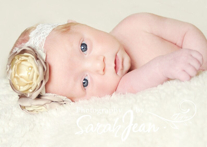 May include: A newborn baby girl wearing a white lace headband with a large beige flower. The baby is lying on a white fluffy blanket. The image is captioned "Photography by Sarah Jean".
