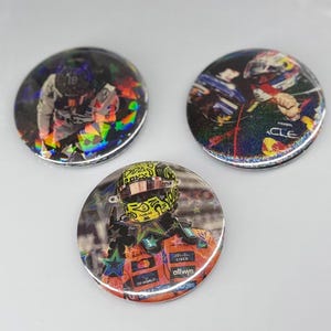 May include: Three circular pin-back buttons featuring holographic star patterns over images of Formula 1 drivers. One button shows a driver in a yellow helmet and orange racing suit, while the others display drivers in racing gear with team logos.