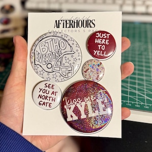 Może przedstawiać: Zestaw pięciu okrągłych przypinek na białej karcie ekspozycyjnej z napisem AFTERHOURS COLLECTORS 5 PACK. Przypinki mają różne wzory, w tym kask futbolowy z napisem BTHO EVERYONE, bordowy tekst JUST HERE TO YELL, SEE YOU AT NORTH GATE, wzór w kowbojki oraz brokatową bordową przypinkę z białym napisem kiss me at KYLE.