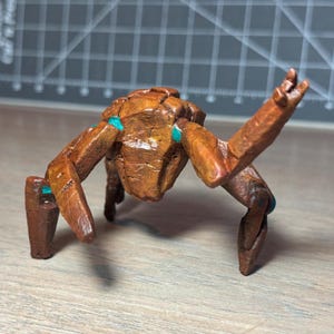 Peut inclure: Une petite figurine en bois sculpté marron représentant une créature à plusieurs pattes avec une surface texturée. La figurine présente des membres articulés et de petits accents bleu sarcelle aux articulations, en position accroupie.