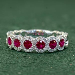 Könnte beinhalten: Ein Silberring mit einer Reihe runder, leuchtend roter Rubine, die jeweils einzeln von einem Kranz aus kleinen, klaren, funkelnden weißen Diamanten umgeben sind. Das Design zeigt ein sich wiederholendes Muster dieser Edelsteingruppen.