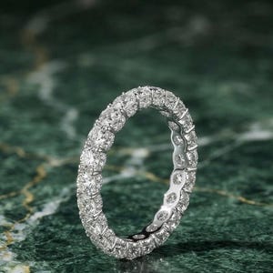 Può includere: Un anello a fascia eternity color argento con una fila continua di pietre trasparenti rotonde a taglio brillante incastonate in una montatura metallica. L'anello è posizionato ad angolo, mostrando la disposizione uniforme delle pietre.